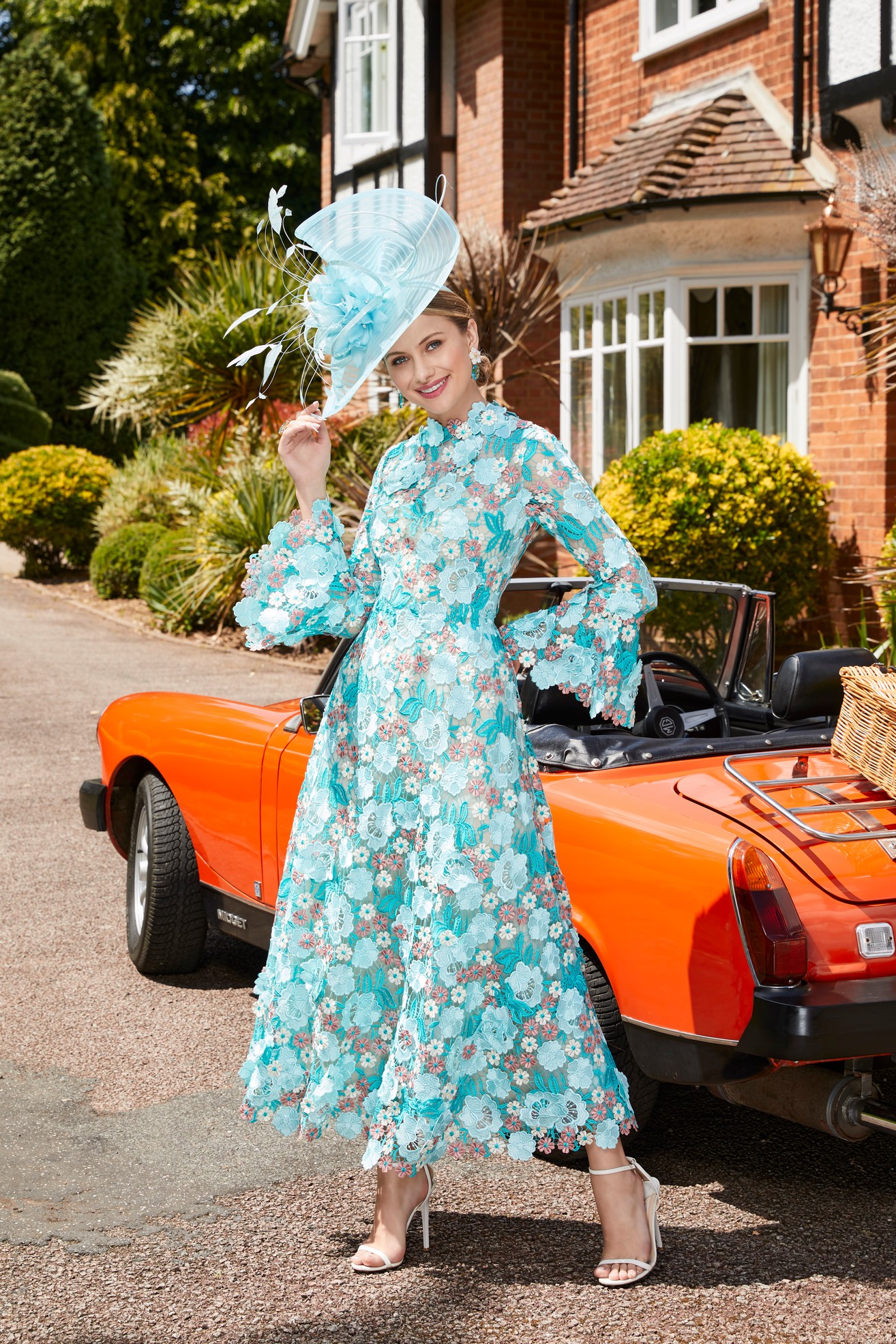 Woman stood next to orange vintage car outside house wearing special occasion a-line jade green dress with high neckline, fluted sleeves and unique 3D floral details 